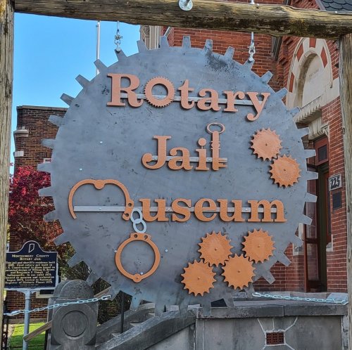 Sign for the Rotary Jail Museum featuring gears and handcuffs, set against a brick building backdrop.
