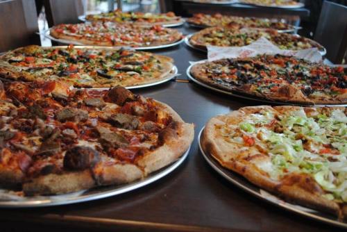 A variety of pizzas on metal trays, featuring different toppings like vegetables, meats, and cheese, on a wooden table.