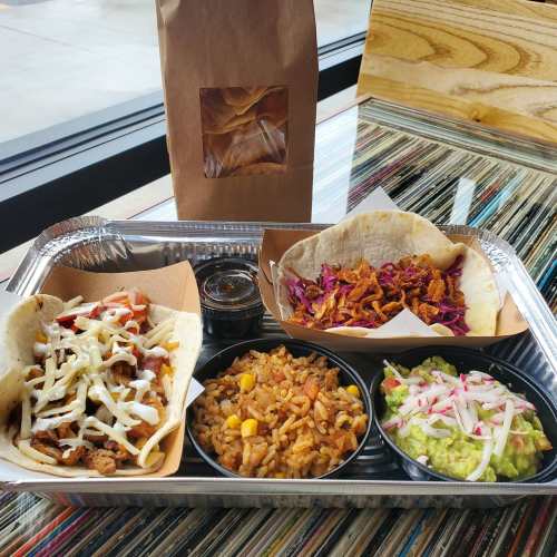A tray with two tacos, rice, guacamole, and a bag of chips on a colorful table.