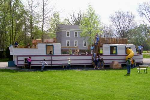 A large, colorful play structure resembling a boat, with children playing on it and nearby grass and trees.