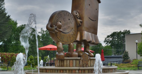 A whimsical bronze statue of a smiling coin character with fountains in a park setting.