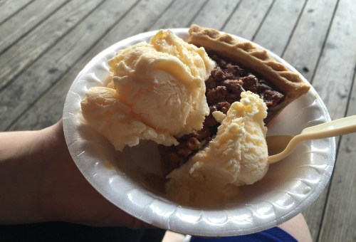 A hand holding a plate with a slice of pie topped with a scoop of vanilla ice cream.