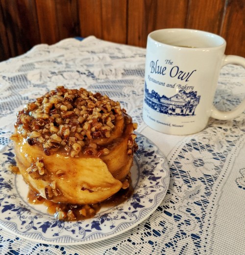 A sticky pecan cinnamon roll on a plate next to a coffee mug from The Blue Owl Restaurant and Bakery.