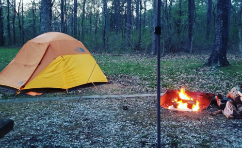 A bright orange and yellow tent set up in a wooded area, with a campfire burning nearby on a gravel path.