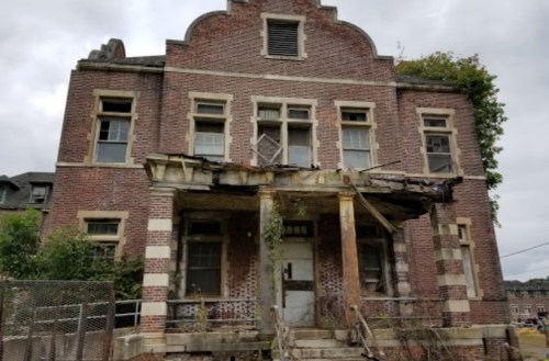 Abandoned brick building with a deteriorating porch and overgrown vegetation under a cloudy sky.