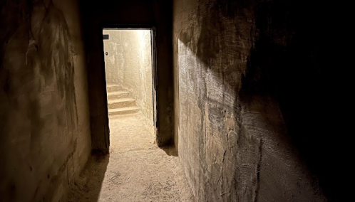 A dimly lit corridor leads to a staircase, with light illuminating the entrance at the far end.