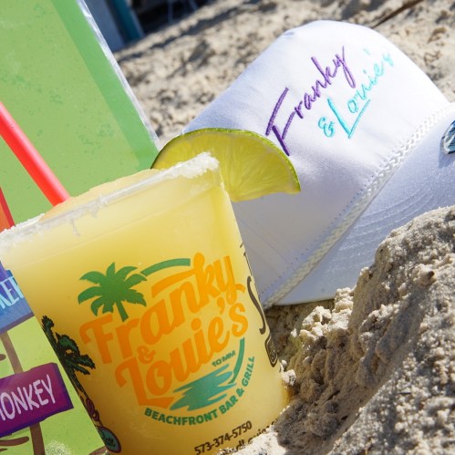 A refreshing drink from Franky & Louie's Beachfront Bar & Grill next to a white cap on the sandy beach.