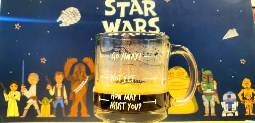 A coffee mug with Star Wars characters in the background, featuring playful text: "Go away! Not yet... How may I assist you?"