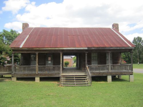 A rustic wooden house with a rusty metal roof, featuring a wide porch and steps, set in a grassy area under a blue sky.