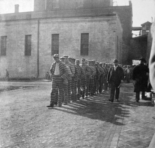 Black and white photo of prisoners in striped uniforms marching in a line outside a building.