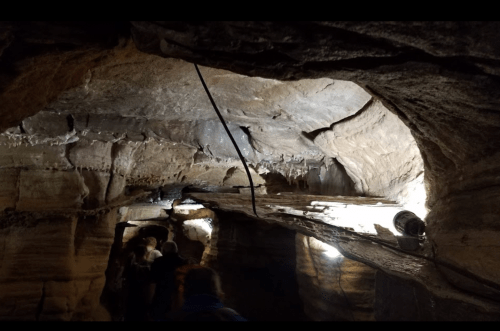 A dimly lit cave interior with rock formations and wooden beams, featuring people walking through the passage.