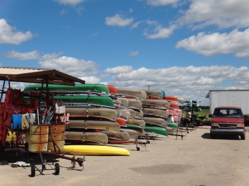 A row of stacked canoes and kayaks under a blue sky with clouds, alongside a parked red vehicle.
