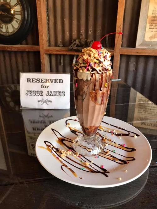 A tall sundae glass filled with chocolate ice cream, topped with whipped cream, nuts, sprinkles, and a cherry, on a plate.
