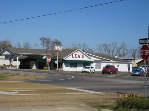 A roadside view of Lea's building with a sign, surrounded by cars and trees, on a sunny day.