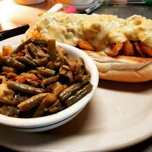 A plate featuring a shrimp sandwich topped with creamy sauce and a side of green beans with bacon.