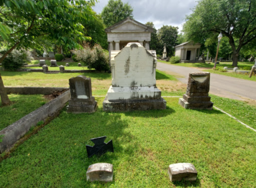 A cemetery scene featuring several gravestones and a large monument surrounded by trees and grass.