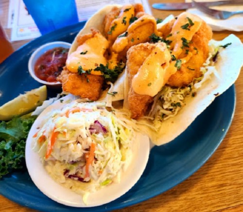 Two crispy fish tacos topped with sauce, served with a side of coleslaw and salsa on a blue plate.