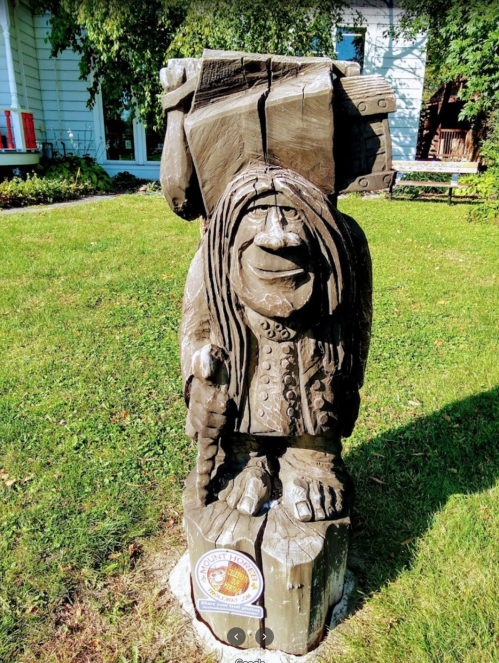 A wooden statue of a smiling figure with long hair, holding a large backpack, set in a grassy area.