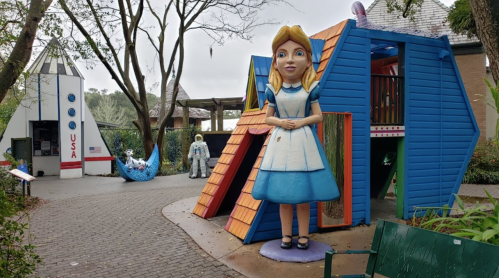 Colorful playground featuring a large Alice figure, a rocket structure, and whimsical play areas surrounded by trees.