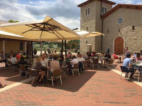 Outdoor dining area with people seated under umbrellas, enjoying meals at tables near a stone building.