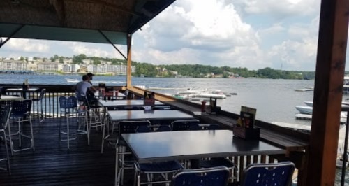 Outdoor dining area overlooking a lake, with empty tables and boats in the water under a partly cloudy sky.