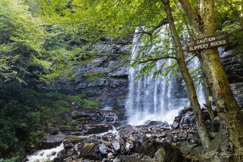 A waterfall cascades down rocky terrain, surrounded by lush green trees and a caution sign for slippery rocks.