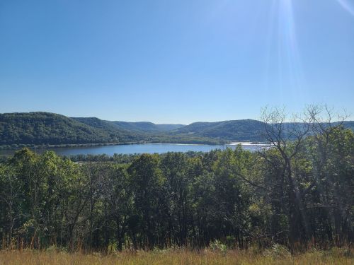A scenic view of a river surrounded by green hills under a clear blue sky. Sunlight glimmers on the water.