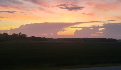 A vibrant sunset with colorful clouds over a green landscape and distant trees.