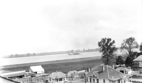 Black and white image of a river with a boat, surrounded by houses and trees along the shoreline.