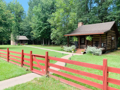 A rustic log cabin surrounded by greenery, with a red wooden fence and a pathway leading to the entrance.