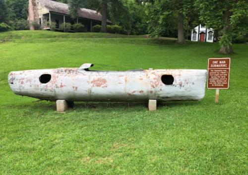 A weathered one-man submarine displayed on grass, with a sign nearby providing information about it.