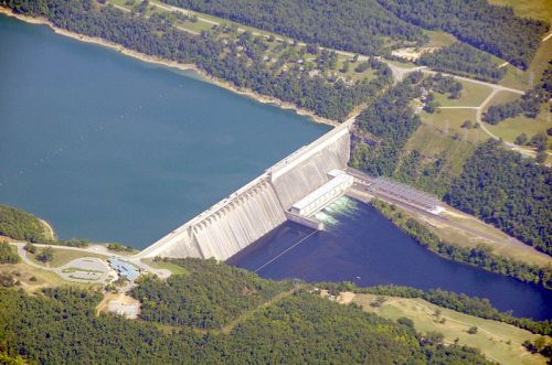 Aerial view of a large dam with water reservoirs on either side, surrounded by green trees and land.