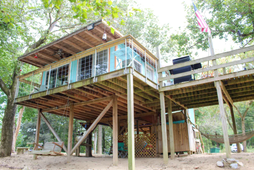 A wooden house elevated on stilts, surrounded by trees, with a deck and an American flag visible.