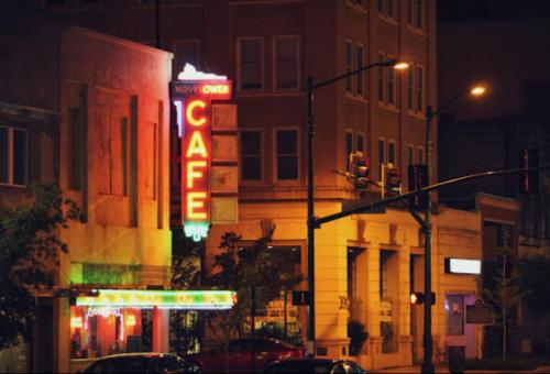 Neon-lit café sign at night, with colorful lights illuminating the street and nearby buildings.