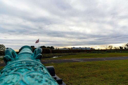 A historic cannon points towards a fort under a cloudy sky, with an American flag waving in the background.