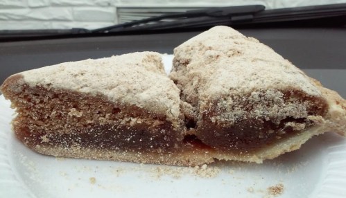 A slice of cake with a crumbly topping, revealing a rich, dark filling inside, served on a paper plate.