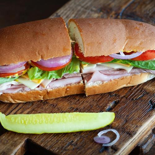 A freshly made sandwich with layers of turkey, lettuce, tomato, onion, and cheese, served with a pickle on the side.