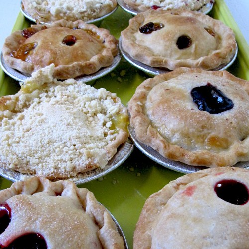 A tray of assorted mini pies with various fillings, some topped with crumble, showcasing golden crusts and vibrant colors.