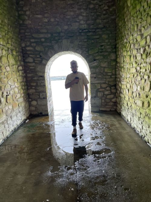 A person stands in a stone archway, surrounded by water, with light shining through the opening behind them.