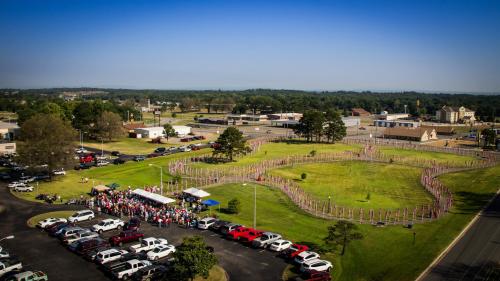 Aerial view of a large crowd gathered in a grassy area surrounded by flags, with buildings and cars in the background.