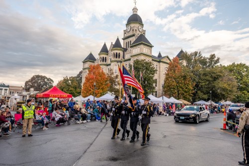 A color guard presents flags in front of a historic building during a parade, with spectators and tents in the background.