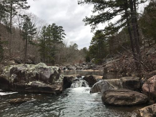 A serene river scene with rocky banks, flowing water, and trees under a cloudy sky.