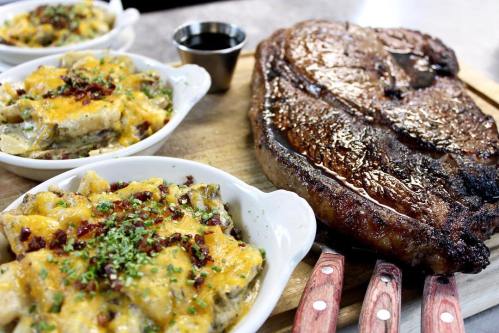 A juicy steak on a wooden board, accompanied by two bowls of cheesy, bacon-topped potatoes.