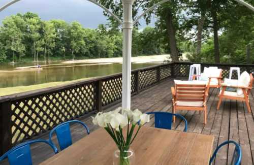 A wooden deck overlooking a calm river, with a table of flowers and outdoor seating under a cloudy sky.