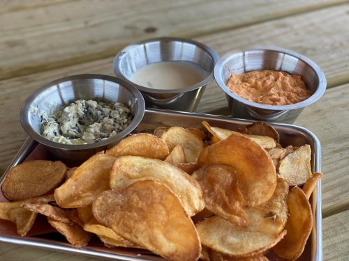 A tray of crispy potato chips served with three dipping sauces in metal bowls: a creamy dip, a spicy dip, and a savory spread.