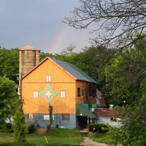 A colorful barn with a quilt pattern on its side, surrounded by greenery and a faint rainbow in the sky.