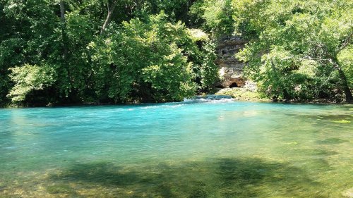 A serene blue river flows through lush green trees, with a rocky cliff in the background.