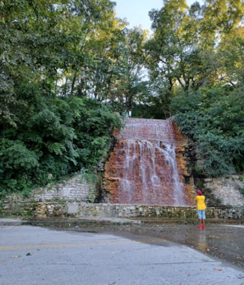 A person stands near a cascading waterfall surrounded by lush greenery and stone walls.