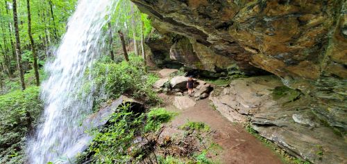 A waterfall cascades over rocks in a lush green forest, with a person standing near the base of the falls.