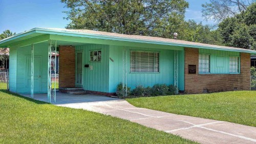 A light blue house with a brick accent, surrounded by green grass and trees, featuring a concrete walkway.